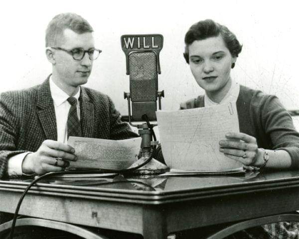 A man and a woman reading from scripts into a microphone with the WILL logo on it