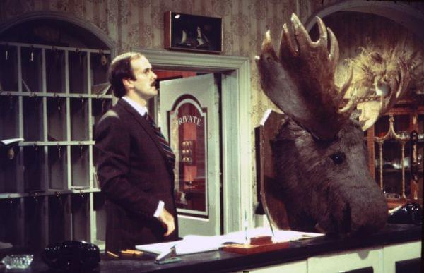 John Cleese behind a hotel desk, with a stuffed moose head nearby.