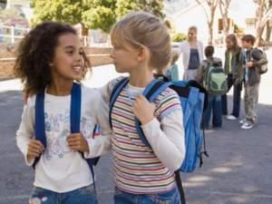 Two young girls with school backpacks look at each other as they walk ahead of a group of classmates