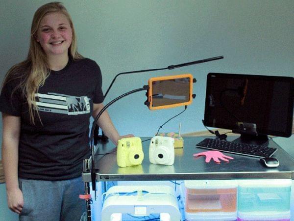17-year-old girl stands next to table with computer keyboard and screen, along with a three-dimensional printer