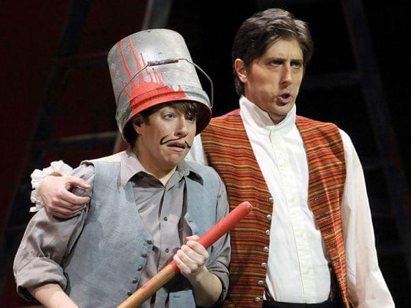 Two people singing onstage, one with a bucket on her head