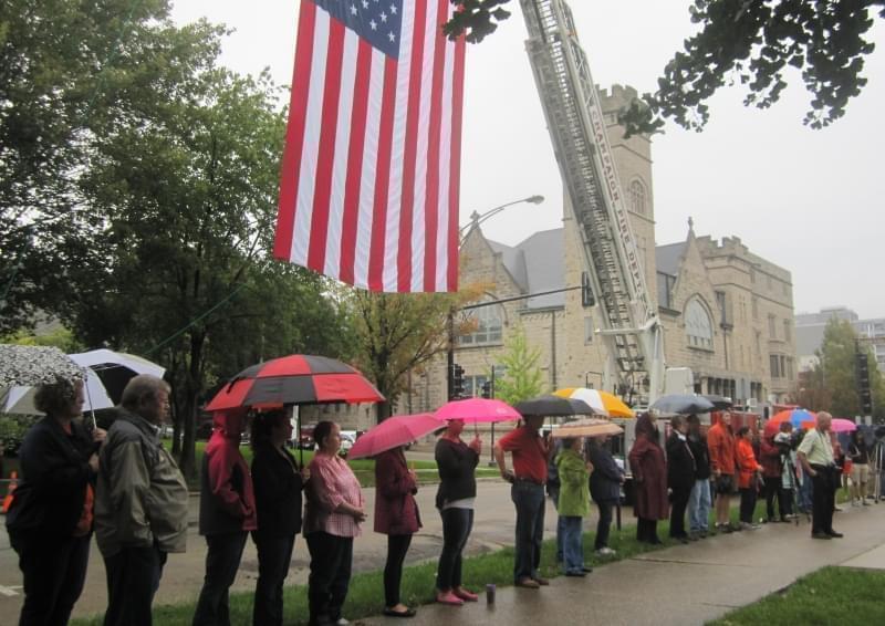 Onlookers with umbrellas gather for Champaign's 9-11 memorial ceremony at West Side Park in 2015, under a US flag hoisted by a crane.