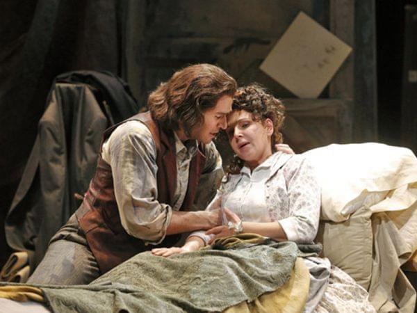 A man sits at the bed side of an ill woman, on stage