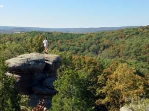 A landscape shot depicting from behind a young woman atop a rock tower looking out over a forest with blue sky beyond.