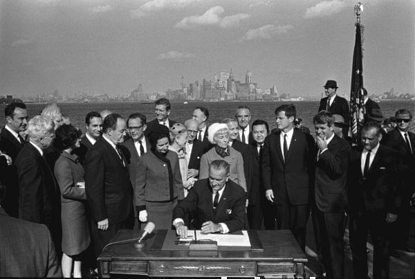 President Lyndon B. Johnson signs the 1965 Immigration and Nationality Act as Vice President Hubert Humphrey, Lady Bird Johnson, Muriel Humphrey, Sen. Edward (Ted) Kennedy, Sen. Robert F. Kennedy, and others look on.