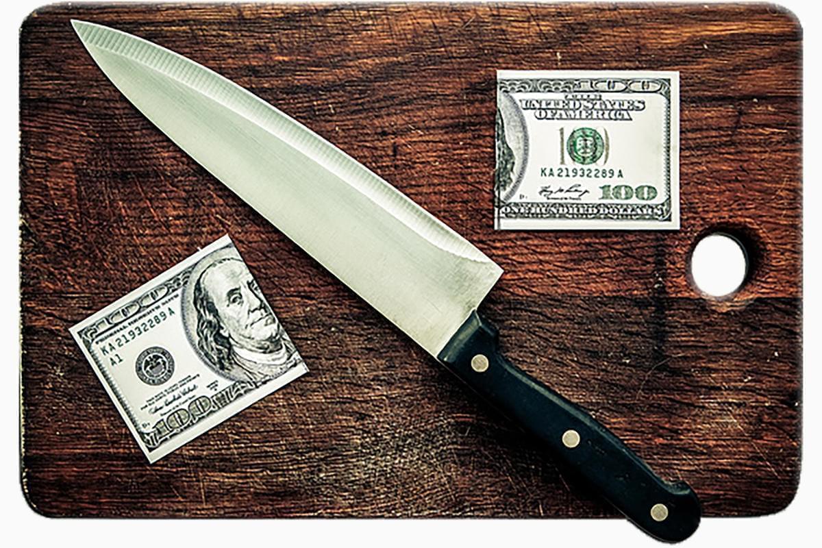 A dollar being cut in half with a knife on a cutting board.