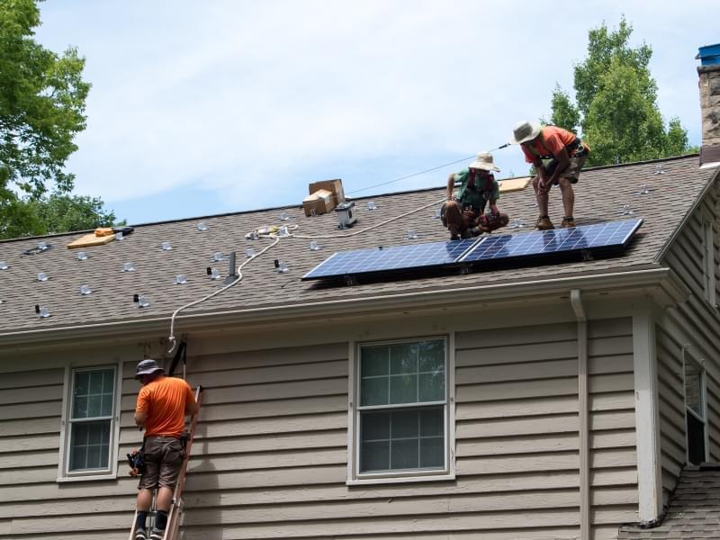 Two men on a roof placing solar panels. Another climbs toward them on a ladder.