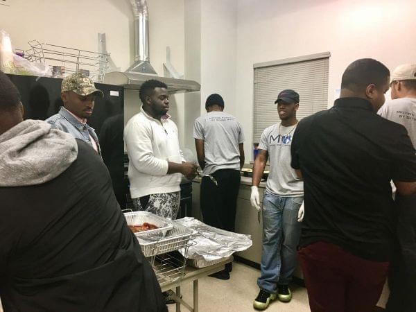 The Men of Impact kitchen crew includes Olivier Jacques (in John Deere hat), Uyi Idemudia (in white hoodie), and Dwayne Mitchell (in the Nike cap).