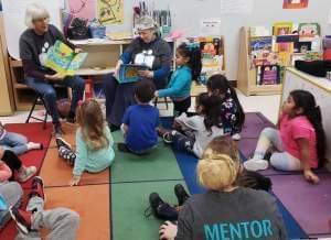 Two adults sitting in chairs in a pre-k classroom reading a book while 3-5 year old children listen while sitting on the rug in front of them.