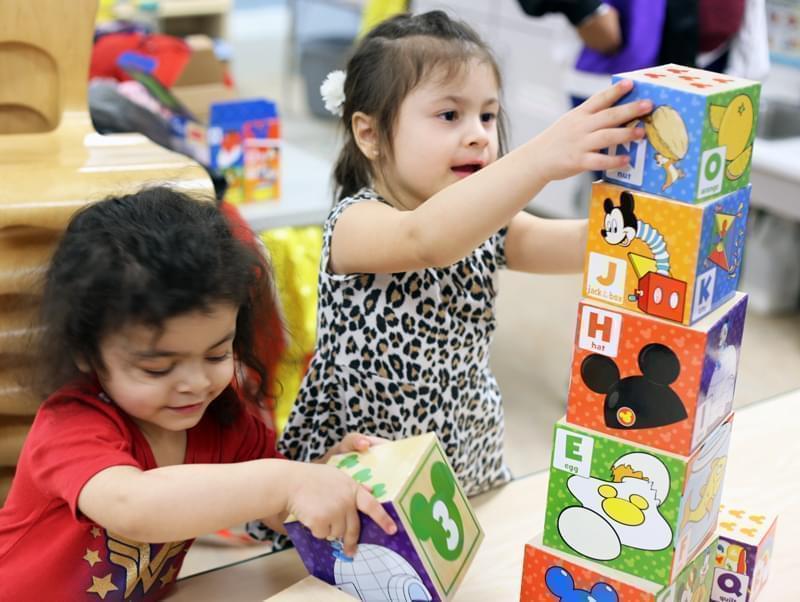 Two 3-year-old children use their hands to build a tower using cardboard nesting blocks.