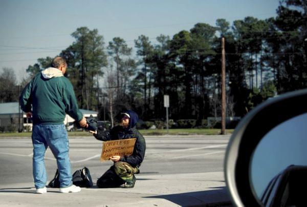 A man asks for money using a cardboard sign that says he is homeless. Another man hands him money while walking by.