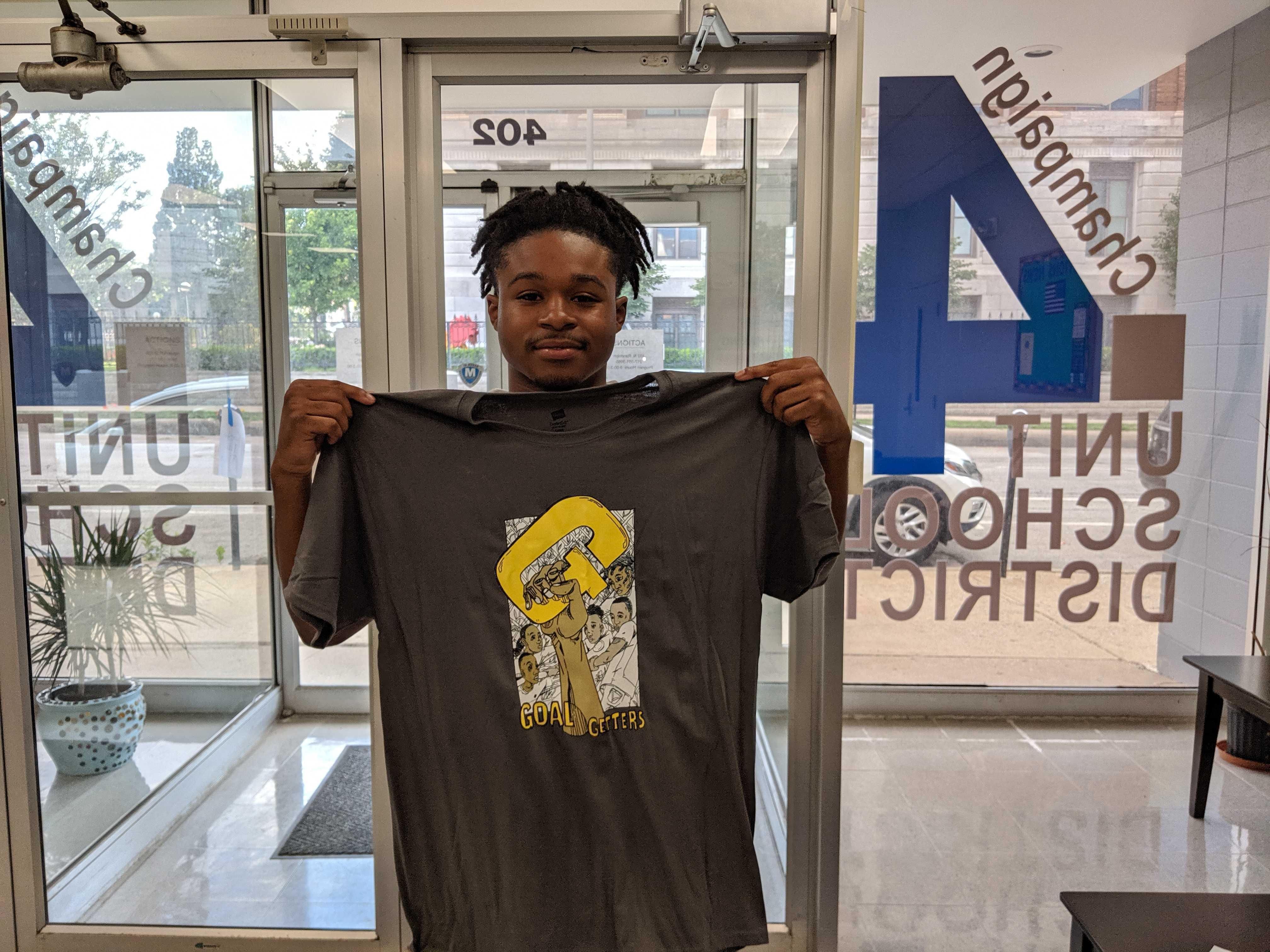Mekel Brown inside a Unit 4 building holding up a Goal Getter T shirt.
