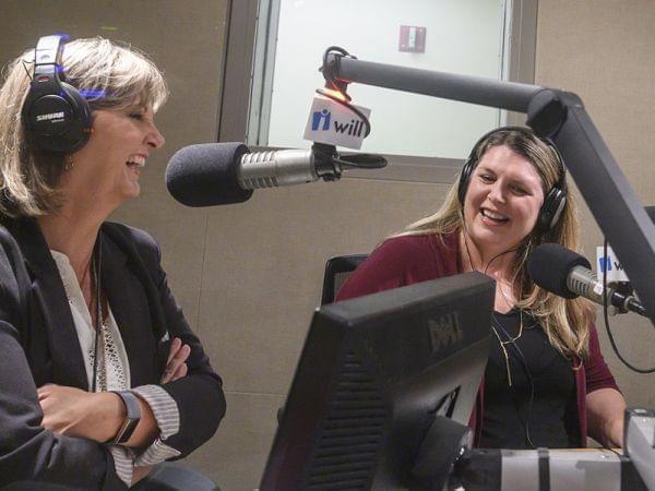 two women sit in a radio studio talking in front of microphones 