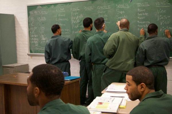 Men in prison stand in front of a black board.