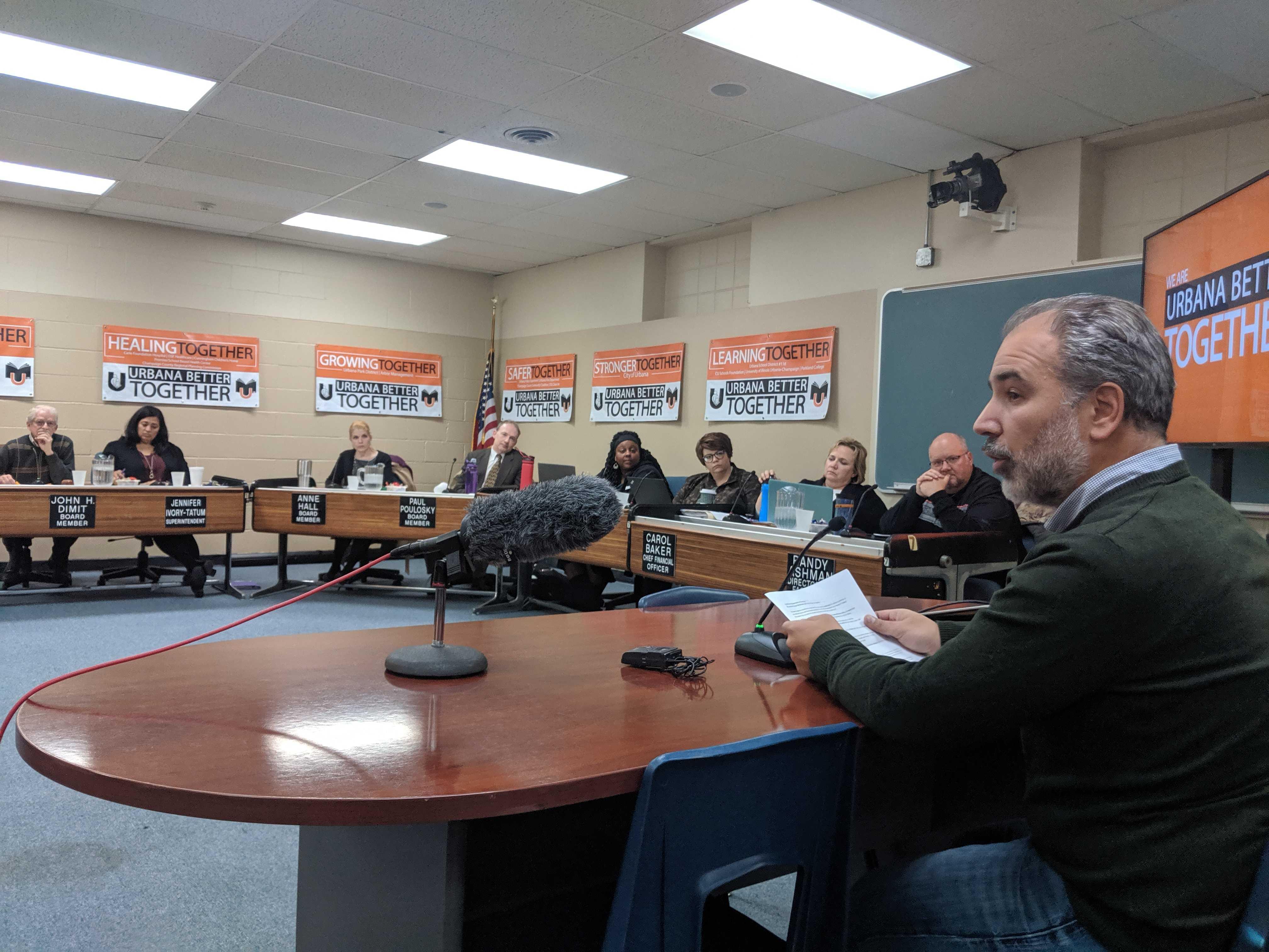 Man sits at a table in front of the board of education in their meeting room to address them during public comment.