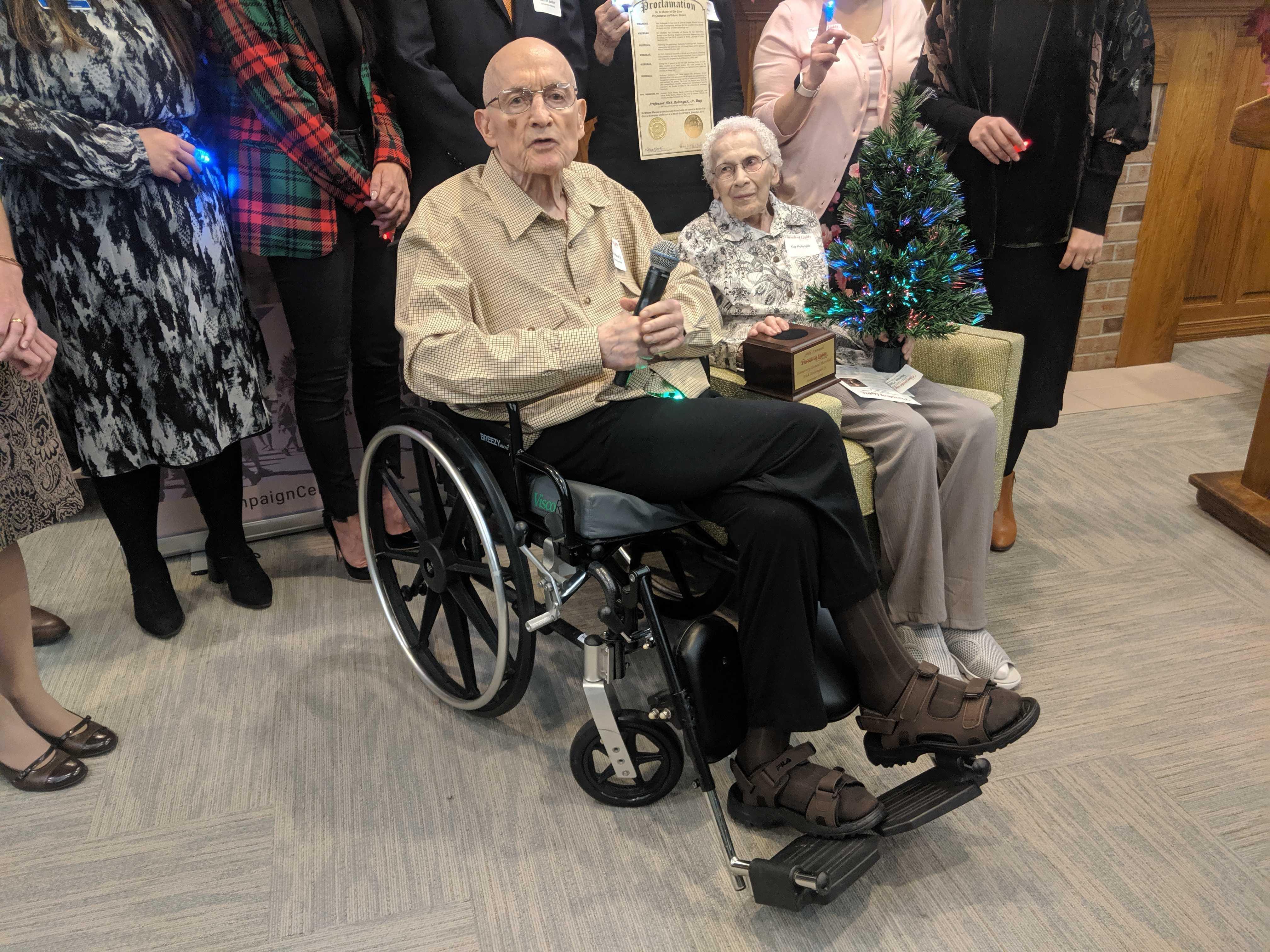 University of Illinois Professor Emeritus Nick Holonyak Jr. and his wife Kay seated with a fiber-optic Christmas tree in front of people standing behind them. 