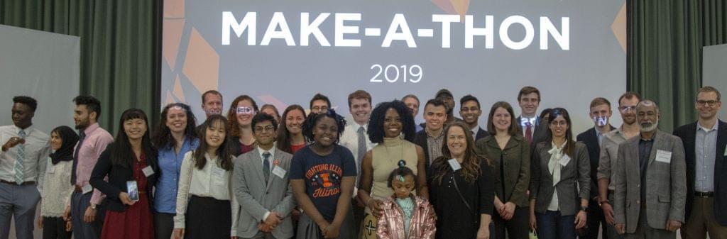 Winners of the first Health Make-A-Thon hosted by the Carle Illinois College of Medicine at the University of Illinois pose with U of I staff. Standing in a row in front a projector screen.