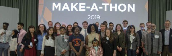 Winners of the first Health Make-A-Thon hosted by the Carle Illinois College of Medicine at the University of Illinois pose with U of I staff. Standing in a row in front a  projector screen. 