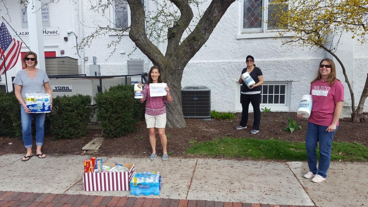 school nurses standing 6 feet apart with cleaning and food supplies outside a homeless shelter