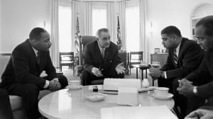 Four men sit around a table in the oval office talking