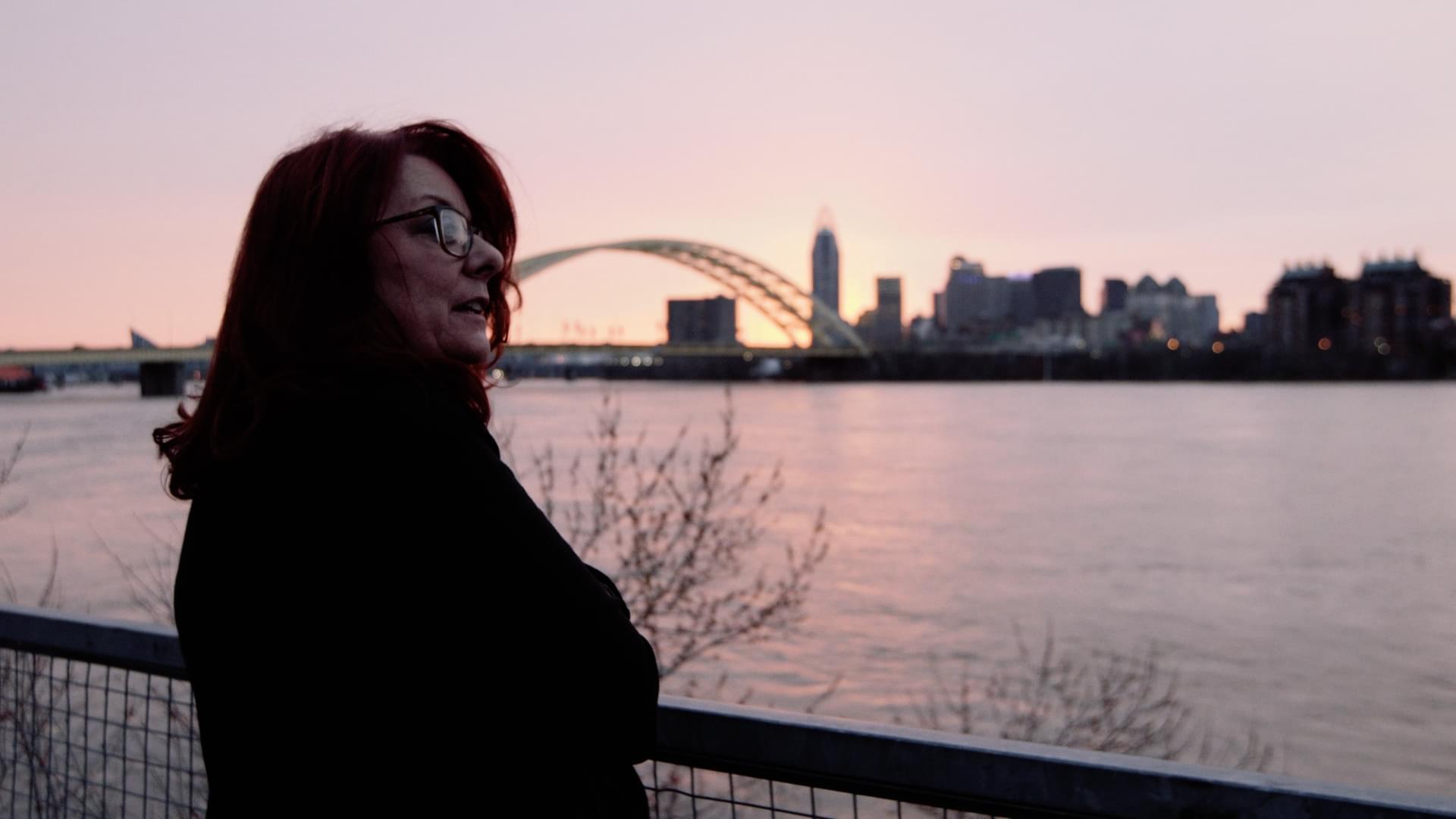 Woman stands next to a river in front of a bridge