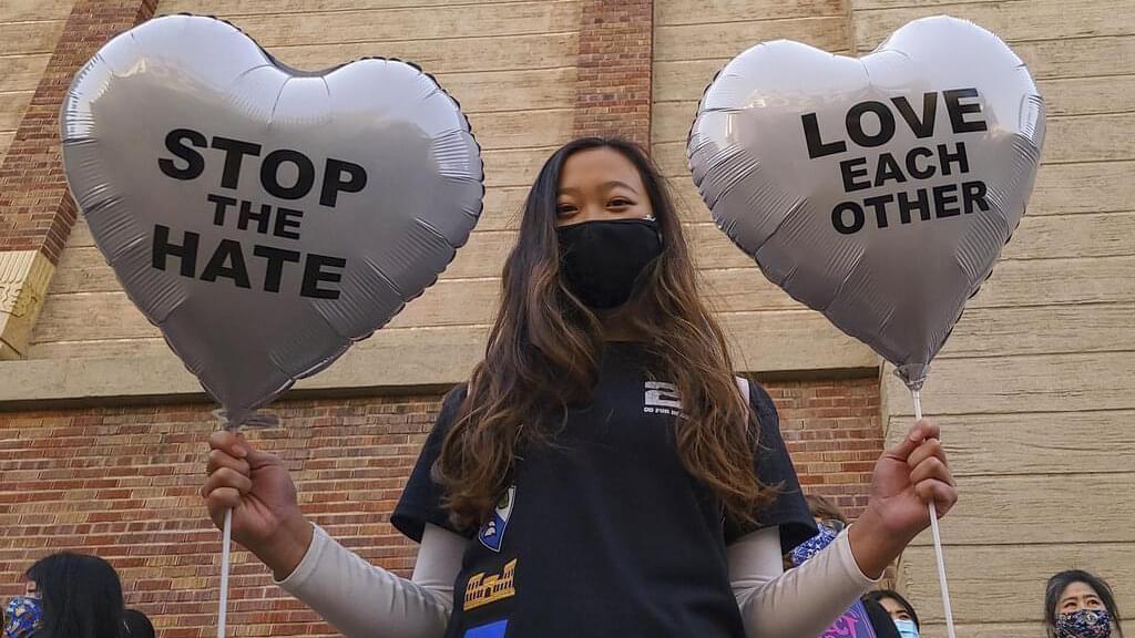 Woman with a mask holds two balloons, one says stop the hate, the other says love each other