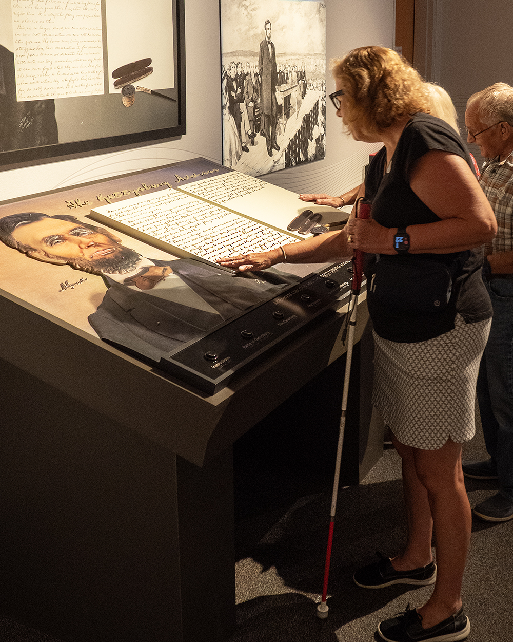 a woman in a back shirt and white skirt holds a red-and-white cane in her left hand while using her right hand to touch a large podium with a 3D replica of the Gettysburg Address
