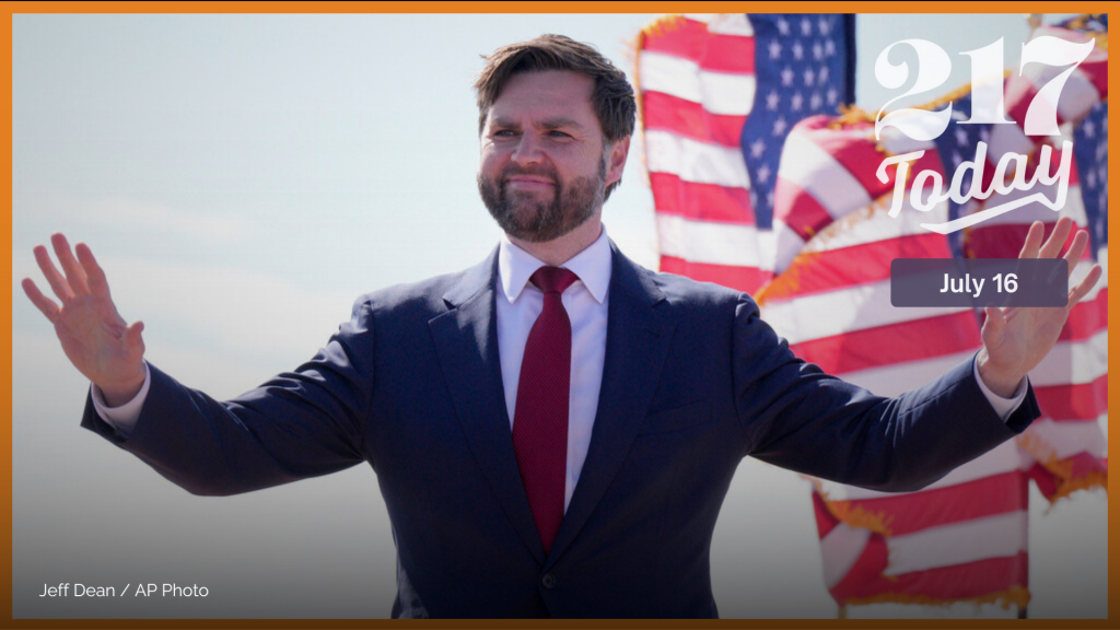 Sen. J.D. Vance has both arms in the air. He waves at an audience while standing in front of a U.S. flag.