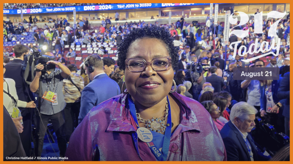 A woman in a purple shirt with glasses smiles at the camera while standing in the United Center for the Democratic National Convention. 