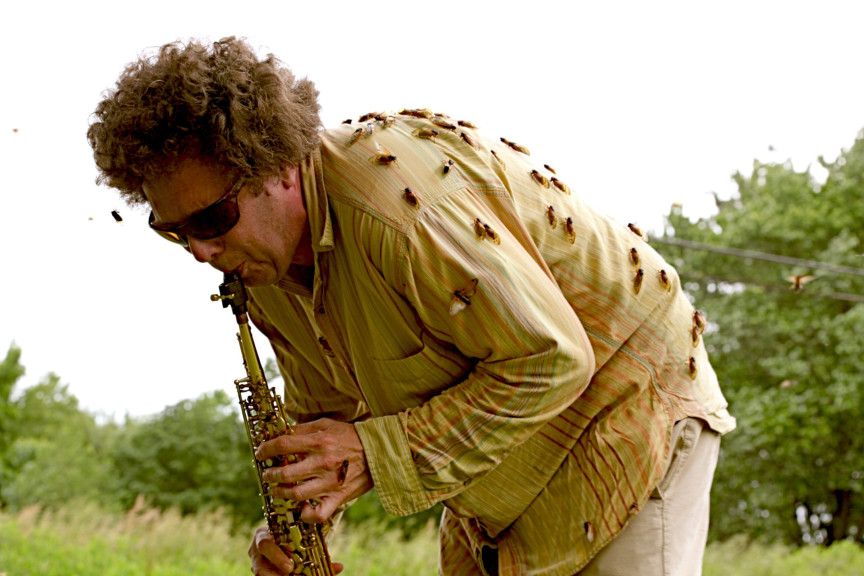 A man with is bent over playing a saxophone with insects covering him.