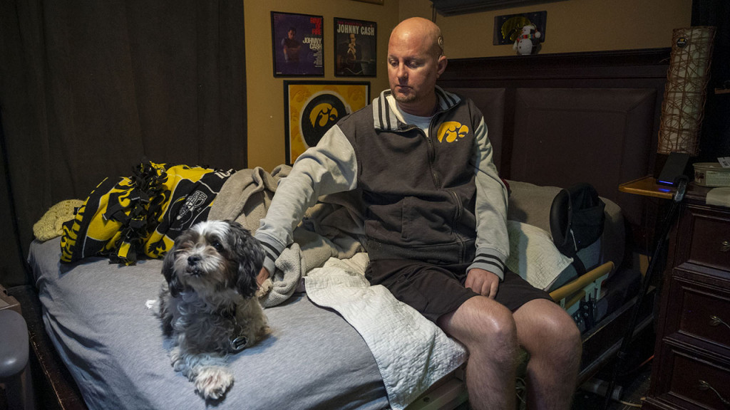  Jon Miller sits in his bedroom with his dog, Carlos, who he received as a present for successfully completing cancer treatment a decade ago. Miller sustained severe brain damage from advanced testicular cancer and its treatment. He requires the help of home health aides to continue living in his West Des Moines home.
