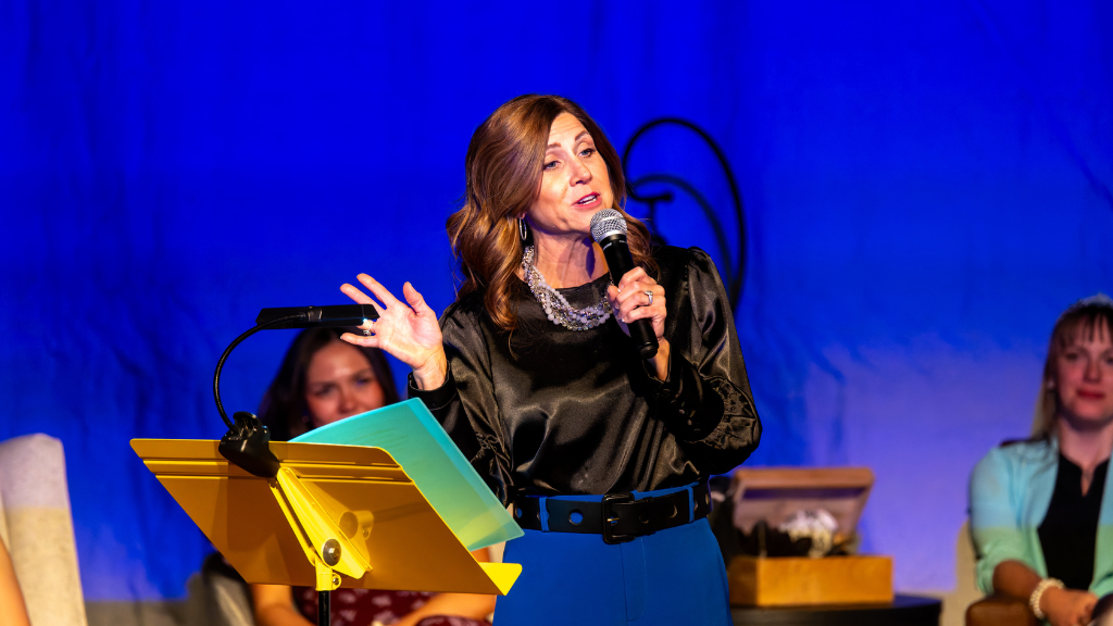 Woman in Black Shirt and Blue pants stands behind yellow podium with microphone in hand. Other women sit behind her. 