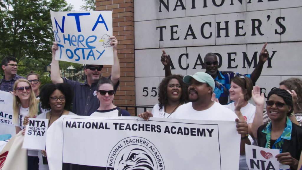 A group of teachers and parents holding a National Teachers Academy banner and protest signs.