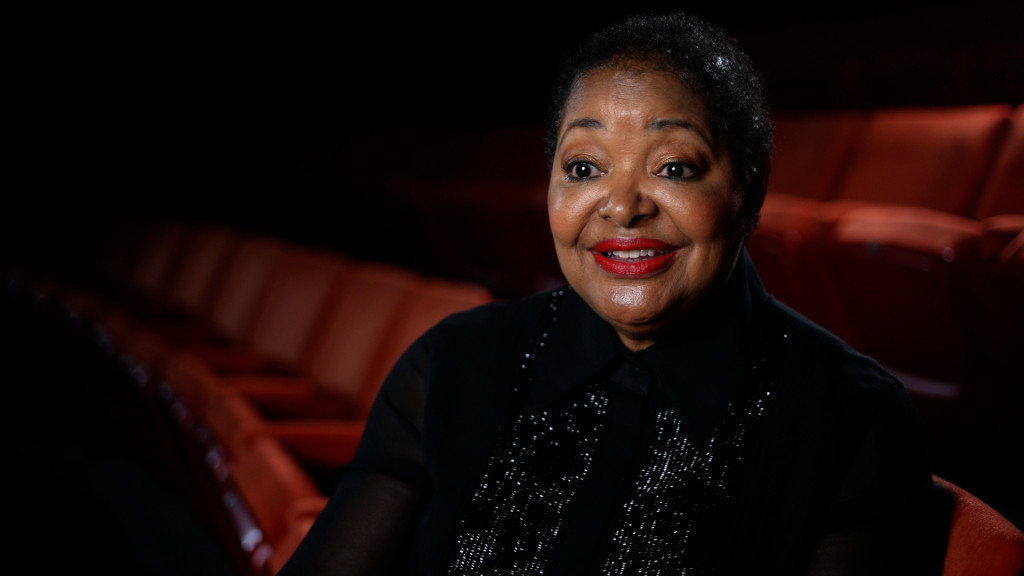 Still from the interview with Dr. Ollie Watts Davis in a theatre with the red theatre seats surrounding Dr. Watts Davis