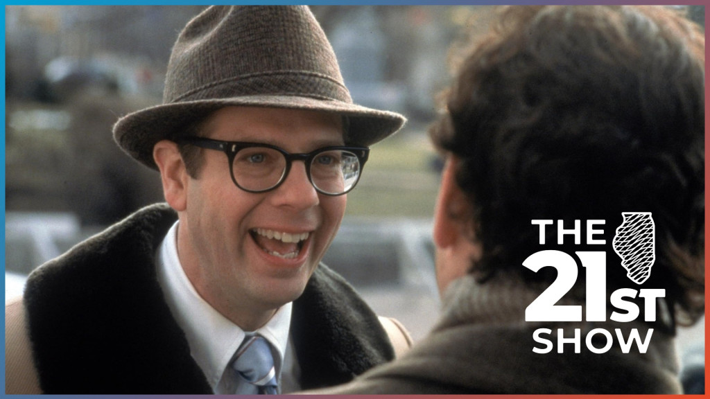 actor Stephen Tobolowsky is seen smiling at Bill Murray, facing away from the camera. Tobolowsky is wearing a fedora, chunky black glasses, and a tan overcoat with wide lapels trimmed in short brown fur