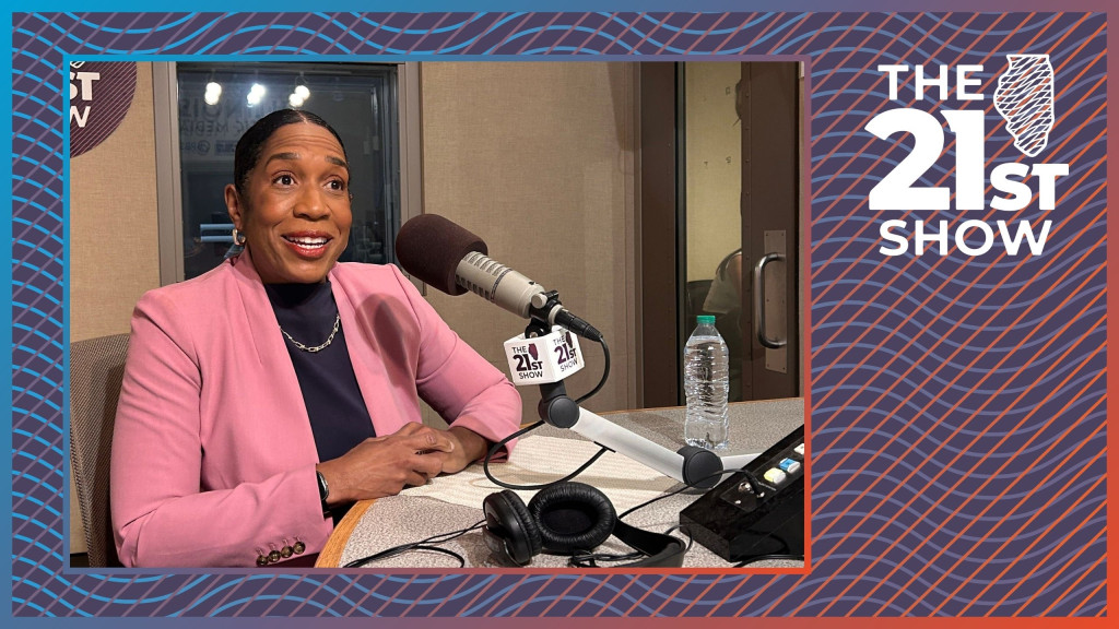 a black woman in a pink blazer and black shirt has her hair pulled back and parted down the middle; she sits in front of a gray Electrovoice RE-20 microphone in radio studio with beige walls