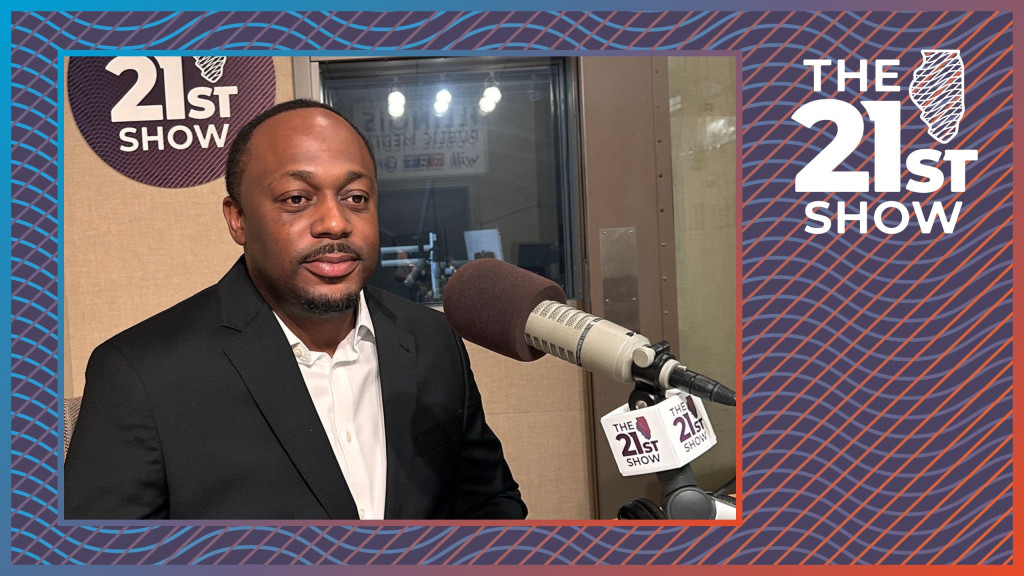 an African-American man wears a dark suit coat and white dress shirt, open at the collar, in a radio studio with a beige background