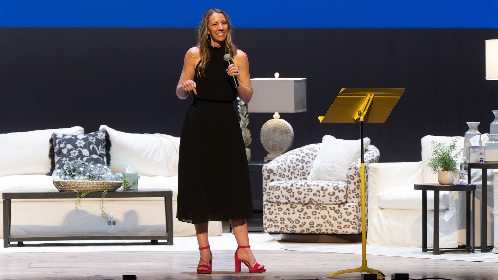 woman in black dress with red heels talks on stage with furniture behind her