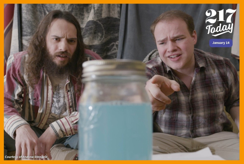 Two men sitting on a couch look at a jar filled with blue liquid.