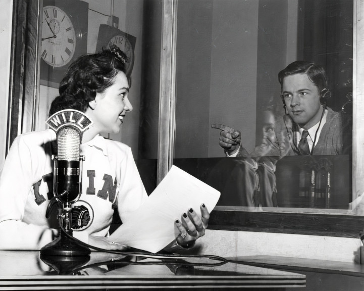 woman sits beyond a microphone looking at man behind glass window
