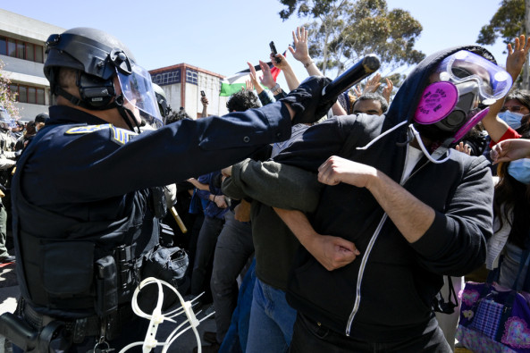 A pro-Palestinian protestor, chained to other protesters with their arms locked, is hit across the face with a baton by a police officer donned in riot gear. 