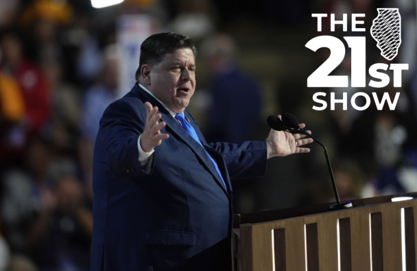 J.B Pritzker wearing a dark blue suit with a bright blue necktie, standing behind a lectern, with his arms open wide