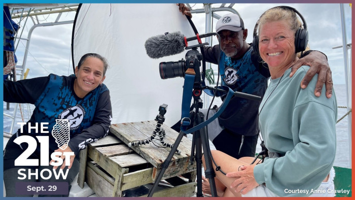 two women and a man smile for a photo on a catamaran sailboat boat during a break from an interview; a wooden crate, multiple cameras on tripods, and the ship's rigging can be seen