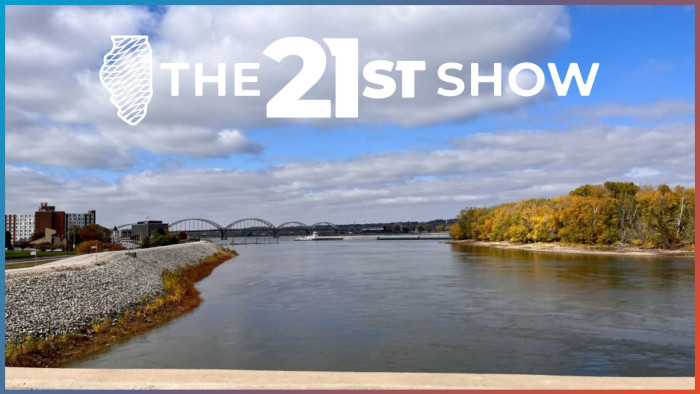 A view of the Mississippi River looking sound. It's is flanked by a path and apartments on the left and a wooded area on the right, where the trees are turning colors in autumn. A bridge over the river is in the distance.