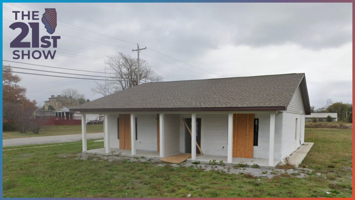 A small white house with plywood over the doors and windows is in a patchy field of grass under overcast skies.