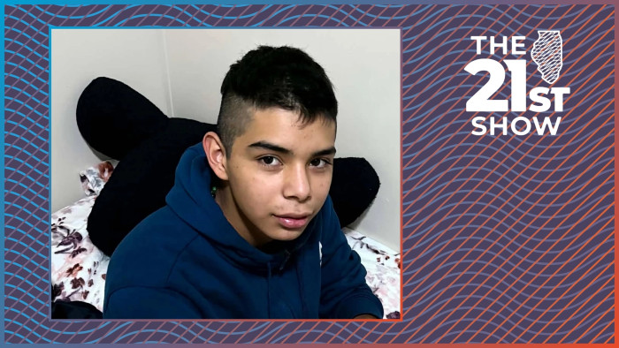 a teenager with dark hair wearing a navy blue hoodie is sitting on a bed