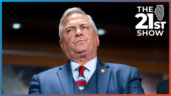 a white man with gray hair has a stern look on his face; he's wearing a dark blue three-piece suit with a light dress shirt and a necktie with red, white and blue stripes