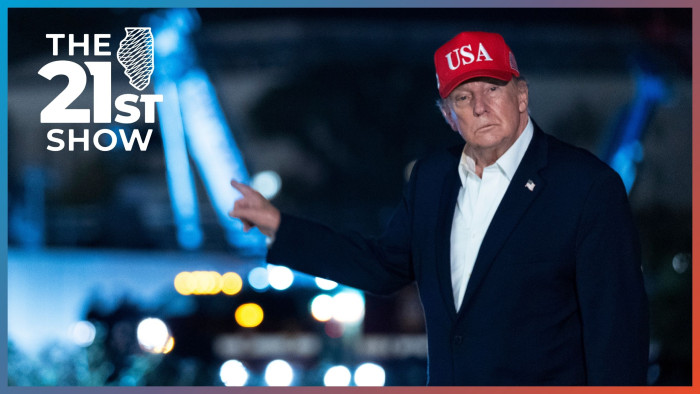 President Trump is wearing a dark-blue suit, white button-down shirt and red USA hat; he looks toward the camera and points behind him; the image was taken at night, so the construction project appears out of focus as a series of color blobs in the background.