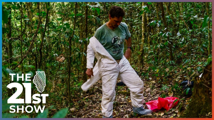 a white man in green T-shirt pulls on a white biohazard suit; there are light blue booties over his shoes and a red biohazard bag lies next to him on a bed of leaves in what appears to be a dense jungle