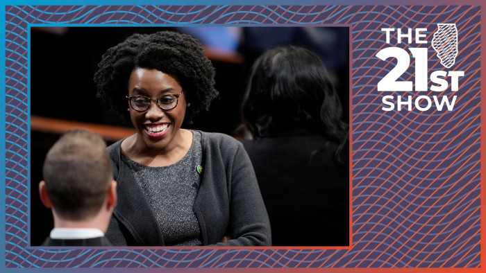 an African American woman in tortoise-shell glasses and a dark charcoal heather sweater smiles as she talks with a white man in a suit facing away from the camera